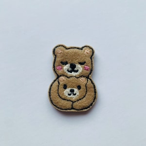 May include: An embroidered patch in shades of brown, depicting two teddy bears embracing. The bears have black eyes, pink cheeks, and black outlines. Suitable for clothing or craft projects.