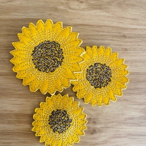 Puede incluir: Tres parches de girasol de fieltro amarillo con centros marrones. Los girasoles son de diferentes tamaños y están dispuestos en un grupo sobre un fondo de madera.