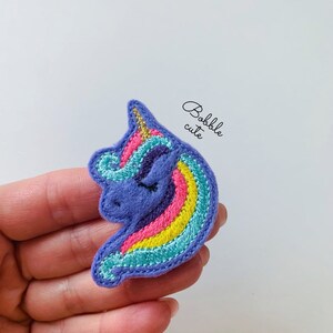 May include: Purple felt unicorn head applique with a rainbow mane and gold horn. The unicorn has closed eyes and a pink nose.
