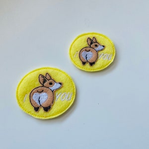 Könnte beinhalten: Zwei gelbe Filzaufnäher mit gestickten Corgi-Hintern-Designs. Einer der Aufnäher sagt "I you" und der andere sagt "I you" mit einem Corgi-Hintern-Design. Die Corgi-Hintern sind braun und weiß, und der Text ist schwarz.