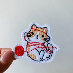 May include: Embroidered iron-on patch featuring a white cat with brown spots and red yarn tangled around it. The cat is holding a ball of red yarn in its paw.
