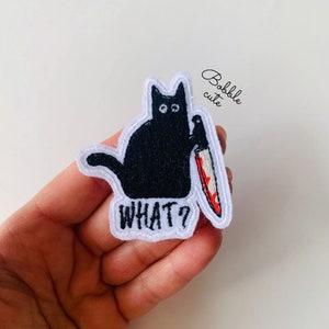 Könnte beinhalten: Schwarze Katze, die ein blutiges Messer hält, mit dem Text "WHAT?" auf einem weißen Patch gestickt.