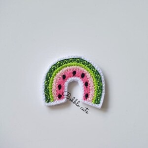 May include: Embroidered patch in the shape of a rainbow, featuring a watermelon design. The rainbow has layers of green, pink, and white, with black seeds. The text "Bobble cute" is written in cursive.