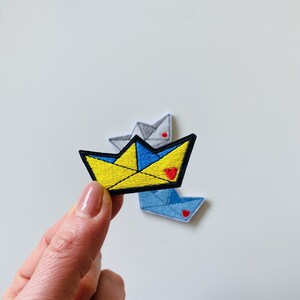 Puede incluir: Parches bordados con forma de barcos de papel. El parche principal es amarillo y azul con un corazón rojo, sostenido por una mano. Se ven dos parches adicionales en gris y azul en el fondo. Los parches tienen bordes negros.