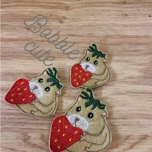 May include: Three embroidered patches featuring a cartoon hamster holding a red strawberry. The hamsters are tan with white faces and black eyes. The strawberries have green tops. The text "Babble cute" is in the upper left corner.