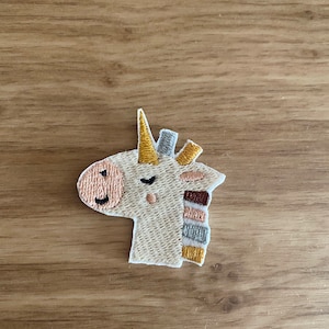 Könnte beinhalten: Ein weißer gestickter Einhorn-Patch mit einem gelben Horn, braunen, rosa und grauen Streifen und einem lächelnden Gesicht.