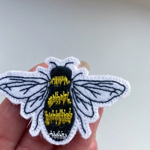 Może przedstawiać: Biała haftowana łatka z pszczołą z czarnymi i żółtymi detalami. Pszczoła ma skrzydła i żądło.