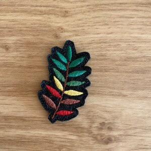 Puede incluir: Un parche bordado con forma de hoja y borde negro. La hoja presenta segmentos verdes, amarillos, rojos y marrones, creando un diseño vibrante y detallado. El parche es ideal para fines decorativos, como adornar ropa o accesorios.