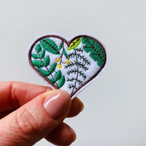 May include: A heart-shaped embroidered patch with a botanical design. The patch features green leaves and yellow accents on a white background, framed by a brown border. The patch is held in a hand.