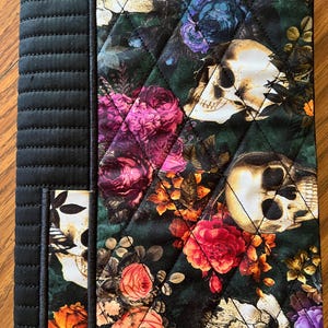 May include: Quilted journal cover featuring a dark floral and skull pattern. The cover has a black spine and a pocket on the left side. The floral design includes roses and other flowers in shades of pink, red, orange, and purple.