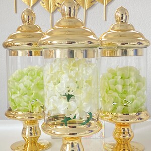 May include: Three decorative glass jars with gold lids and bases. The jars contain green and white flowers. The jars are on a white surface, with gold feather-shaped decor in the background.