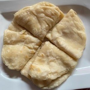 Puede incluir: Vista cenital de varios trozos de pan plano cocido de color marrón dorado, dispuestos sobre un plato blanco. El pan plano parece suave y ligeramente hinchado, con texturas y pliegues visibles. Los bordes muestran un ligero dorado.