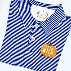 May include: A blue and white horizontally striped polo shirt with a collar and two white buttons. An embroidered orange pumpkin with a brown stem and the white monogram letters BB is featured on the chest. A white tag is tucked into the collar.