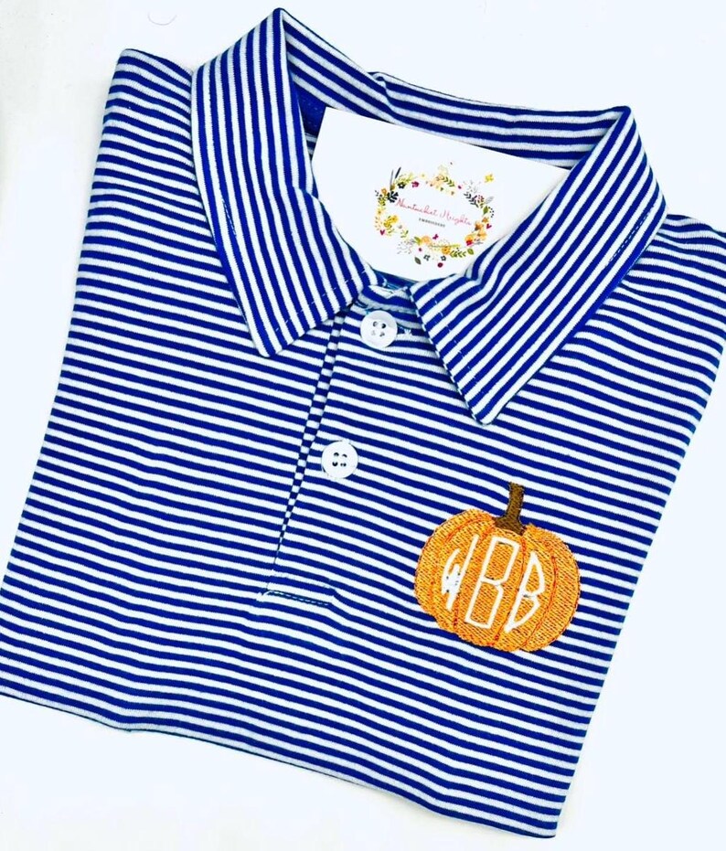 May include: A blue and white horizontally striped polo shirt with a collar and two white buttons. An embroidered orange pumpkin with a brown stem and the white monogram letters BB is featured on the chest. A white tag is tucked into the collar.