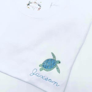 May include: White t-shirt with an embroidered sea turtle and the name "Jaxson" in blue thread.