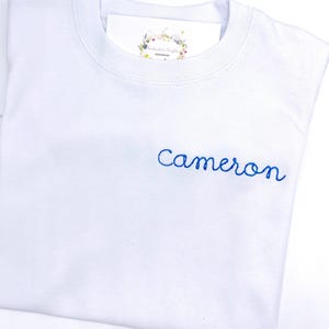May include: A white crew neck sweatshirt with the name "Cameron" embroidered in blue thread on the front. A small card with floral illustrations is tucked into the neckline.