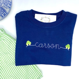 May include: A navy blue t-shirt with the name "carson" embroidered in white thread. Two small green cars with yellow wheels are embroidered on either side of the name.