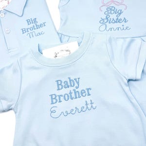May include: Light blue baby clothing set with embroidered text. Includes a polo shirt with "Big Brother Mac", a top with "Big Sister Annie", and a onesie with "Baby Brother Everett".