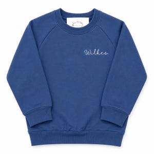 May include: A solid navy blue long-sleeve sweatshirt with a crew neckline and ribbed cuffs and hem. The word Wilkes is embroidered in white cursive script on the upper left chest. A white brand label is visible inside the collar area.
