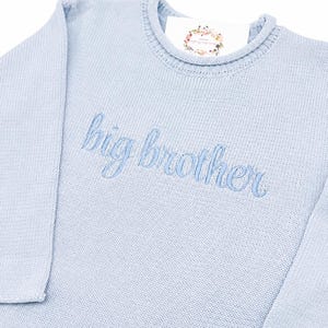 May include: A light blue long-sleeve knit jumper featuring the embroidered text "big brother" in a matching blue script font across the chest. The garment has a ribbed crew neckline and includes a white brand tag with a floral wreath graphic.