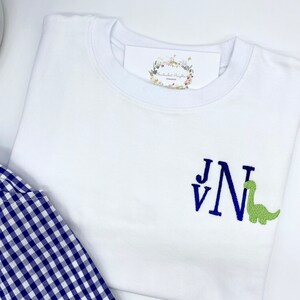 May include: White t-shirt with a green dinosaur and blue embroidered initials "J V N".