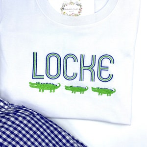 May include: A white short-sleeved t-shirt featuring the embroidered name LOCKE in blue and green outlined letters, positioned above three small green embroidered alligators. A navy and white gingham patterned fabric is visible at the bottom.