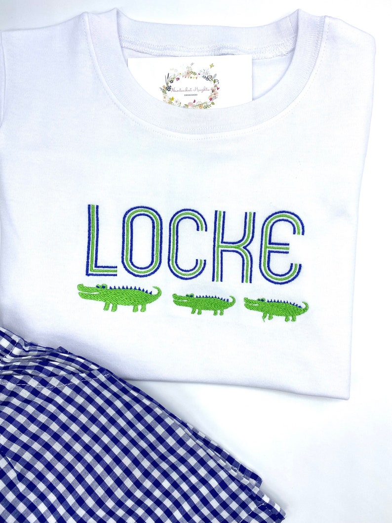 May include: A white short-sleeved t-shirt featuring the embroidered name LOCKE in blue and green outlined letters, positioned above three small green embroidered alligators. A navy and white gingham patterned fabric is visible at the bottom.