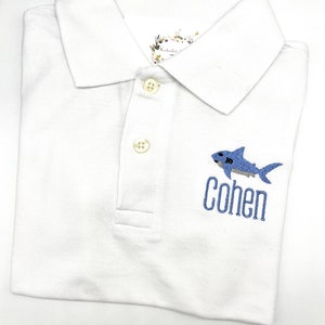 May include: White polo shirt with a blue embroidered shark and the name "Cohen" underneath.