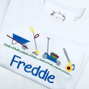 May include: White t-shirt with blue embroidered text "Freddie" and a design of a wheelbarrow, a rake, and a lawnmower on a green grass background.