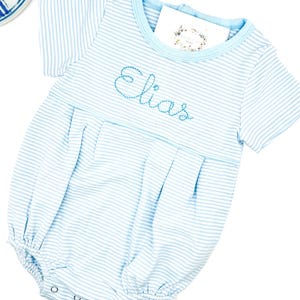 May include: Light blue and white striped baby romper with the name "Elias" embroidered in blue script. The romper has short sleeves, a round neckline, and snap closures at the bottom. A small card with floral design is tucked into the neckline.