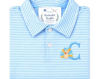 May include: A light blue and white striped polo shirt featuring a collar and two white buttons. An embroidered letter C is on the right side, decorated with a colorful turkey design. A white tag reads Nantucket Heights Embroidery.