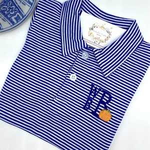 May include: A blue and white striped polo shirt featuring a collar, two white buttons, and an embroidered monogram with the letters WRB above a small orange pumpkin. A white tag with floral graphics and text is tucked inside the collar area.