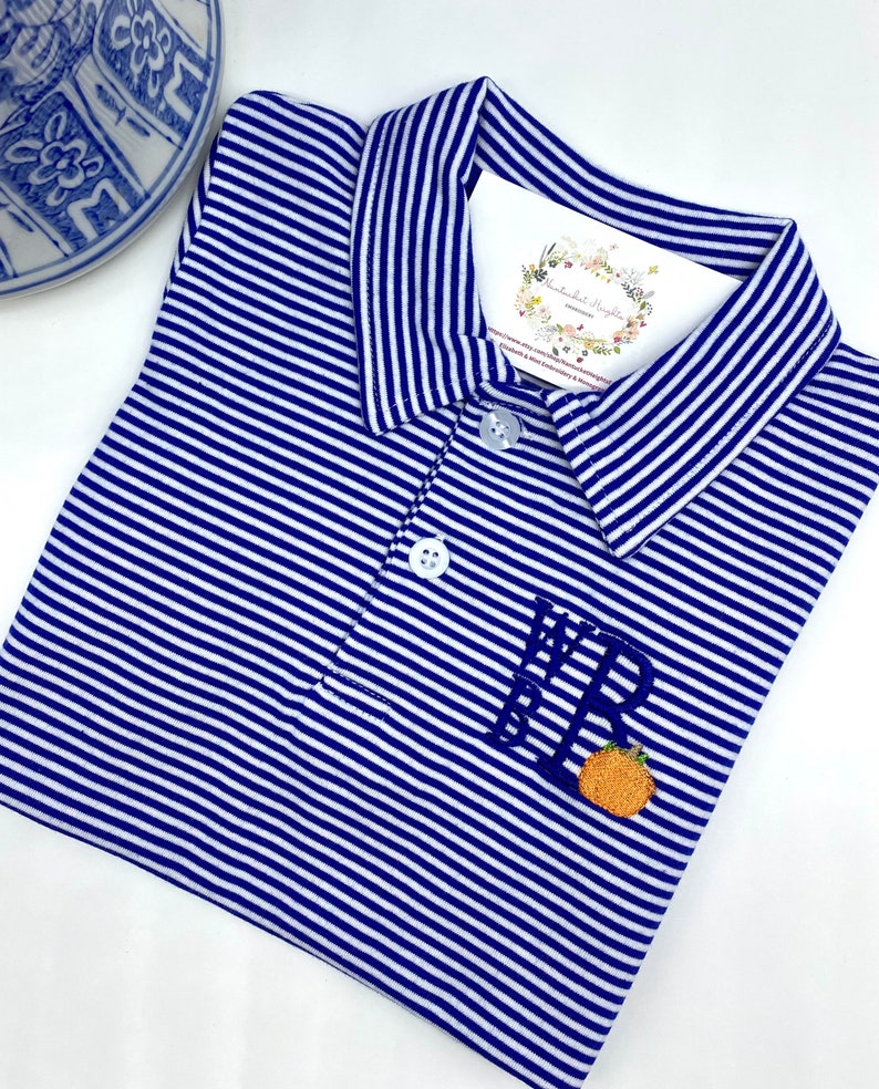 May include: A blue and white striped polo shirt featuring a collar, two white buttons, and an embroidered monogram with the letters WRB above a small orange pumpkin. A white tag with floral graphics and text is tucked inside the collar area.