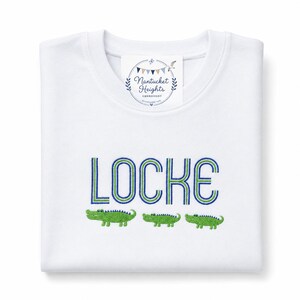 May include: A white long-sleeve shirt featuring the embroidered name LOCKE in blue and green block letters. Below the name are three small green embroidered alligators. A white tag at the neckline displays the text Nantucket Heights Embroidery.