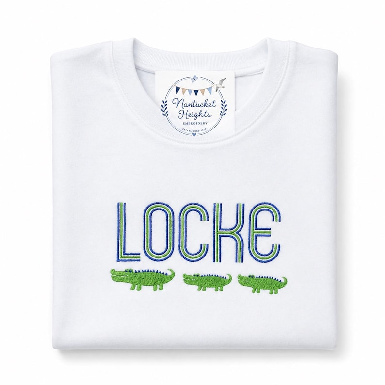 May include: A white long-sleeve shirt featuring the embroidered name LOCKE in blue and green block letters. Below the name are three small green embroidered alligators. A white tag at the neckline displays the text Nantucket Heights Embroidery.