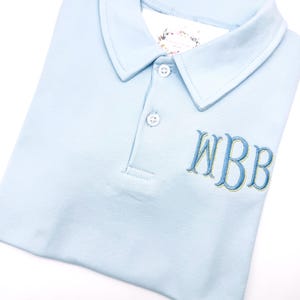 May include: A light blue polo shirt with a three-button placket and a collared neckline. The shirt features the embroidered letters "WBB" in a contrasting blue color. The shirt is folded neatly on a white surface.