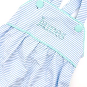 May include: Light blue and white striped baby romper with the name "James" embroidered in light blue thread. The romper has light blue buttons and trim. The straps are light blue and white striped.