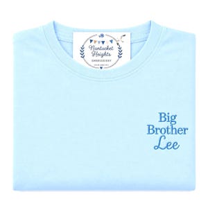 May include: A light blue t-shirt featuring dark blue embroidered text that reads Big Brother Lee on the lower right side. A white rectangular tag is placed at the neckline with the text Nantucket Heights Embroidery and a graphic of bunting.