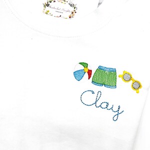 May include: White t-shirt with embroidered beach-themed designs. Features a beach ball, green swim trunks, and yellow sunglasses. The name "Clay" is embroidered in blue script. A floral label is visible.