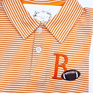 May include: Orange and white striped polo shirt with a white collar. The shirt features an embroidered orange letter "B" and a brown football. The shirt has two white buttons.