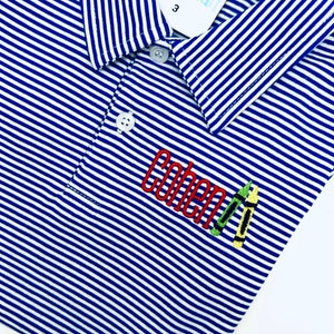 May include: A blue and white striped polo shirt featuring a collar, two white buttons, and an embroidered design on the chest. The embroidery displays the name Cohen in red thread, accompanied by two small crayon illustrations in green and yellow.