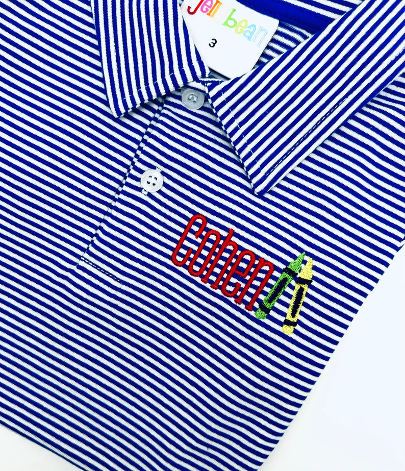 May include: A blue and white striped polo shirt featuring a collar, two white buttons, and an embroidered design on the chest. The embroidery displays the name Cohen in red thread, accompanied by two small crayon illustrations in green and yellow.
