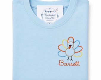 May include: A light blue t-shirt featuring an embroidered turkey design on the lower right side with the name Barrett stitched in orange thread below it. A white tag with the text Nantucket Heights Embroidery is visible at the neckline.
