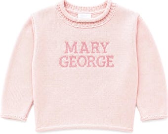 Może przedstawiać: Jasnoróżowy sweter z dzianiny z długim rękawem, wykończony ściągaczem przy okrągłym dekolcie i mankietach. Napis MARY GEORGE jest wpleciony w nieco ciemniejszym odcieniu różu na środku klatki piersiowej, tworząc spójną fakturę.