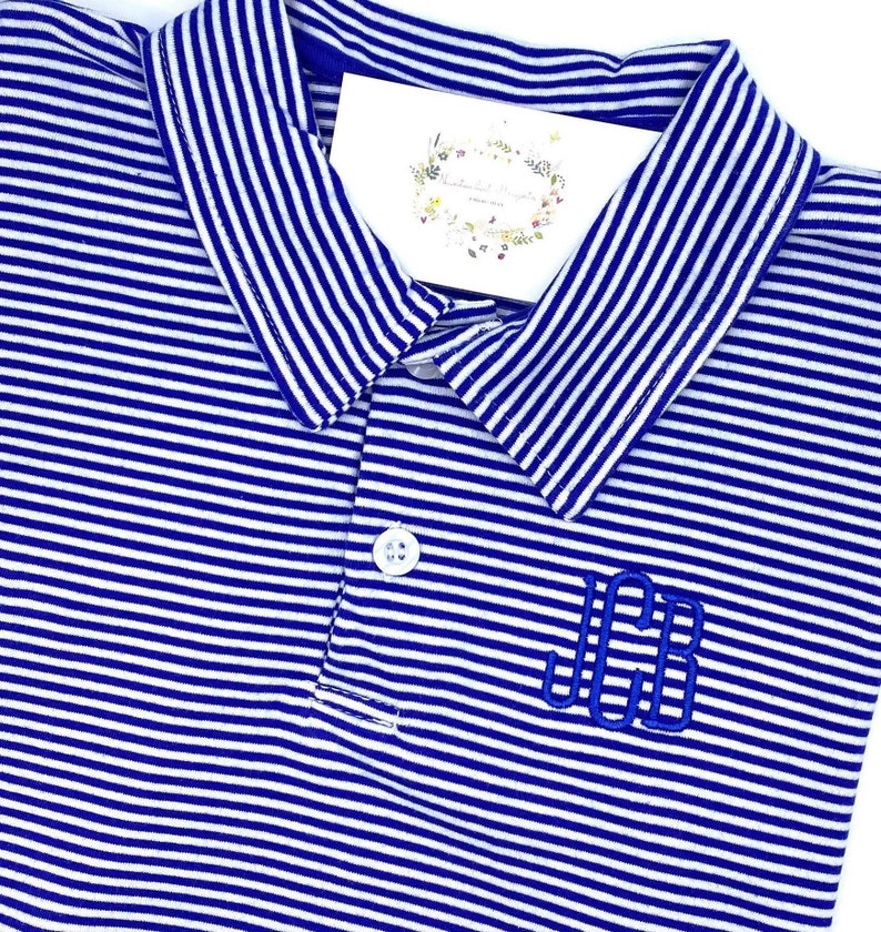 May include: A blue and white striped polo shirt featuring a classic collar, a single white button at the placket, and the embroidered monogram letters JCB in blue thread on the chest. A small white tag with floral graphics is tucked inside.