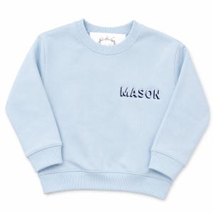 May include: A light blue long-sleeved sweatshirt featuring a ribbed crew neckline, cuffs, and hem. The name MASON is embroidered in dark blue capital letters on the right side of the chest. A white brand label is visible inside the collar.