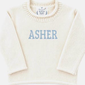May include: A cream-coloured long-sleeve knit jumper featuring a ribbed crew neckline and the name ASHER embroidered in light blue block letters across the chest. A white label inside the collar reads Nantucket Heights.