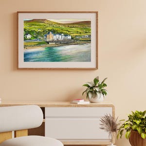 Könnte beinhalten: Ein gerahmtes Aquarellgemälde einer Küstenlandschaft. Das Kunstwerk zeigt einen Strand, Gebäude und grüne Hügel unter einem weichen, farbenfrohen Himmel. Der Rahmen besteht aus hellbraunem Holz.