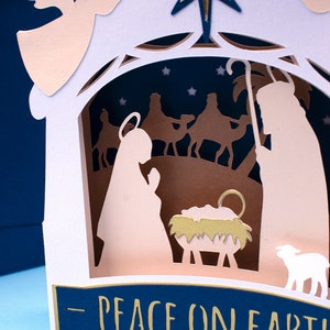 May include: A white paper Christmas card with a pop-up nativity scene. The scene features Mary, Joseph, a baby Jesus in a manger, a sheep, and three wise men riding camels. The card has a blue background with gold stars and the text "Peace on Earth".