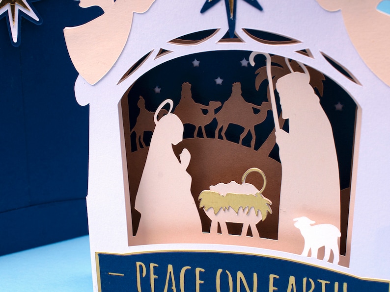 May include: A white paper Christmas card with a pop-up nativity scene. The scene features Mary, Joseph, a baby Jesus in a manger, a sheep, and three wise men riding camels. The card has a blue background with gold stars and the text "Peace on Earth".